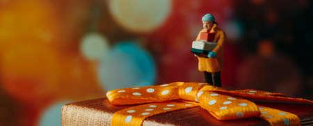 closeup of a miniature man, wearing a warming coat, carrying a pile of boxes standing on top of a gift box in a bokeh effect background, in a panoramic format to use as web banner or headerの写真素材