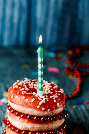 closeup of a lit candle on top of a pile of donuts covered with a red frosting, on a gray rustic wooden tableの写真素材
