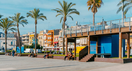 Tarragona, Spain - December 29, 2021: People enjoying the sun on a winter day sitting on the benches of the promenade by the sea at El Serrallo, the traditional fishermen district in Tarragona, Spainのeditorial素材