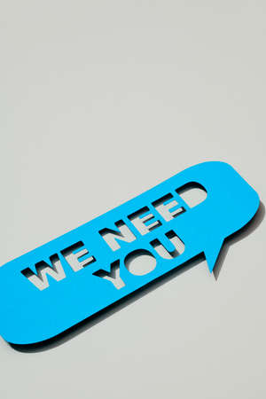 closeup of a blue paper speech bubble, with the text we need you cut out on it, on a gray background with some blank space on topの写真素材
