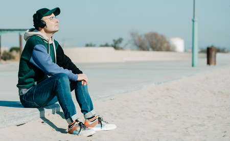 a middle-aged man, wearing a multicolored hoodie and jeans, listens to music using a pair of headphones sitting on the promenade, next to the sand of a urban beach on a sunny winter dayの写真素材