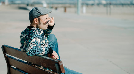 closeup of a middle-aged man, wearing colorful modern casual clothes, sitting in a bench outdoors, in a urban space, looking over his sunglassesの写真素材