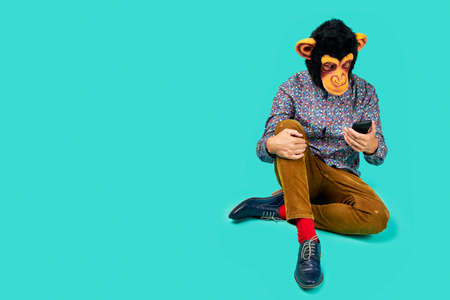 a young man wearing a monkey mask and colorful clothes, sitting on the floor in a blue background with some blank space on the left, is watching his smartphone, as having a video callの写真素材