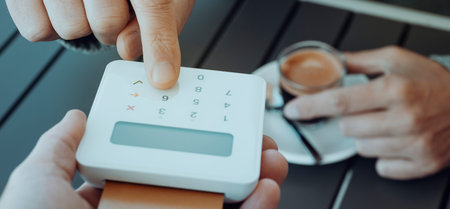 a young caucasian man, sitting on an outdoors table, inserting the PIN of his credit card on the...