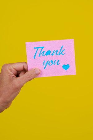 a young caucasian man has a pink piece of paper with the text thank you written in blue, on a yellow backgroundの写真素材