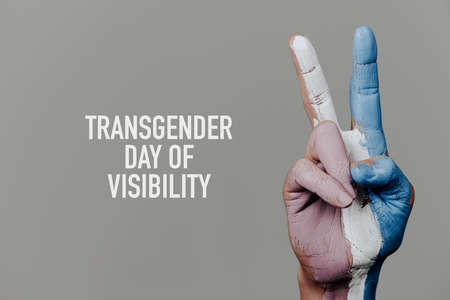 the text transgender day of visibility and the hand of a person doing the V-sign with the transgender pride flag painted in it, on a pale gray backgroundの写真素材