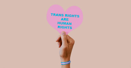 a young caucasian person, wearing a wristband patterned with the transgender pride flag, holds a...