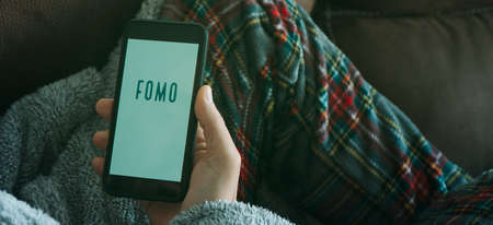 closeup of a young man in the sofa, wearing pajamas and house robe, holding his smartphone, that reads the text fomo, for fear of missing out, in his hand, in a panoramic format to use as web bannerの写真素材