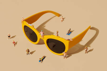 some miniature people, wearing swimsuit, relaxing on top of a pair of yellow plastic-rimmed sunglasses and some more miniature people standing around, against a pale brown backgroundの写真素材
