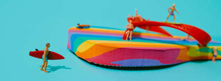 closeup of a miniature man carrying a red surfboard next to a rainbow-patterned flip-flop, with some other miniature men in swimsuit, on a blue background, in a panoramic format to use as web bannerの写真素材