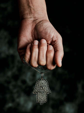 closeup the hand of a man holding an old hamsa amulet, also known of the hand of fatima or the hand of mary, on a black mottled backgroundの写真素材