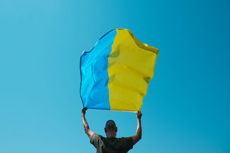 a man, seen from behind, waves the ukrainian flag above his head on the sky in a sunny dayの写真素材