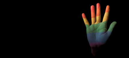 the hand of a person, patterned with the rainbow pride flag, emerges from the black background, with some blank space on the left, in a panoramic format to use as web banner or headerの写真素材