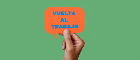 man holds an orange paper speech bubble with the text back to work written in spanish, on a green...