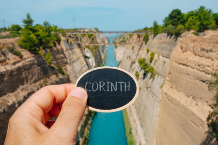 closeup of a man holding a black sign with the text Corinth written in it, in front of the Corinth...
