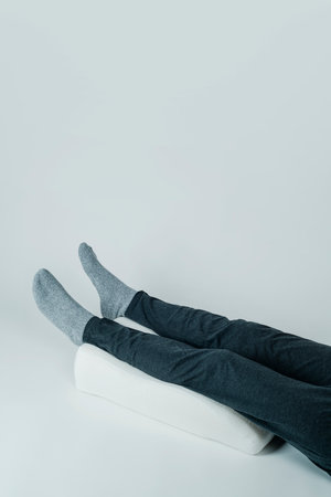 a man uses a white leg elevation pillow, made of memory foam, on an off-white background with some blank space on topの写真素材