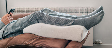 a man leans his legs on a leg elevation pillow made of memory foam, resting on the sofa, in a...