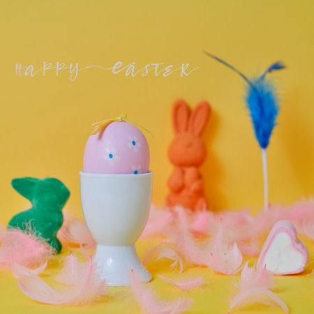 text happy easter and a pink easter egg in a white eggcup, some pink feathers and some easter bunnies of different colors on a yellow backgroundの写真素材