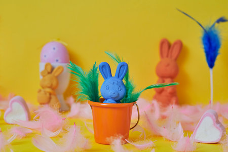 a blue easter bunny in an orange bucket surrounded by pink feathers and a pink easter egg and some other easter bunnies of different colors on a yellow backgroundの写真素材