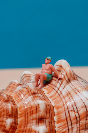 closeup of a miniature man, in a green swimsuit, wearing a green cap and a pair of sunglasses, sitting on a seashell, against a blue background with some blank space on topの写真素材