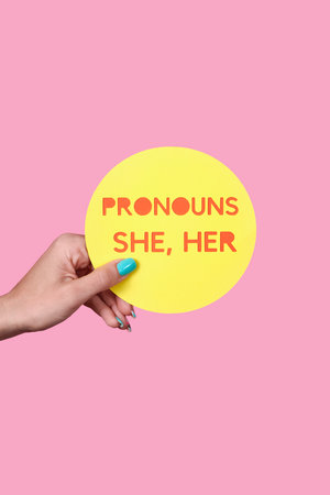 a caucasian person holds a yellow round sign with the text my pronouns are she, her in front of a pink backgroundの写真素材