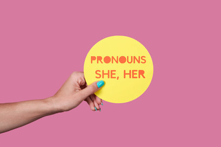 closeup of a caucasian person holding a yellow sign with the text my pronouns are she, her on a pink backgroundの写真素材