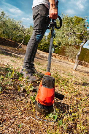 closeup of a young man mowing the grown grass of a farmland using a cordless electric string trimmer