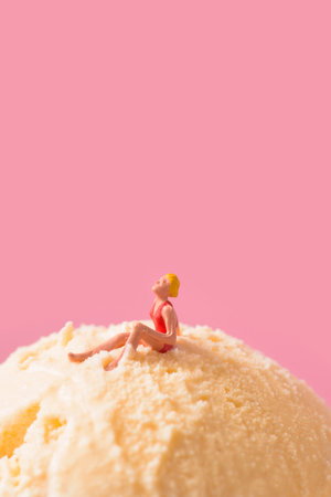a miniature woman, wearing a red swimsuit, sitting on top of a vanilla ice cream ball, against a pink background with some blank space on topの写真素材
