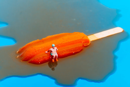closeup of a miniature man wearing a green swimsuit, a green swimming cap and  swimming goggles sitting on a melting orange ice pop on a blue backgroundの写真素材