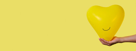 a man holds a smiley yellow heart-shaped balloon in his hand on a yellow background with some blank space on the left, in a panoramic format to use as web bannerの写真素材
