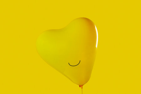 closeup of an inflated yellow heart-shaped balloon with a smile drawn in it on a yellow background, to depict happinessの写真素材