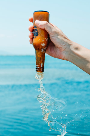 a man holds an open beer bottle upside-down in front of the water, on a summer dayの写真素材
