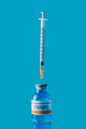 a simulated vial of fentanyl with a syringe in its rubber stopper on a blue surface against a blue backgroundの写真素材