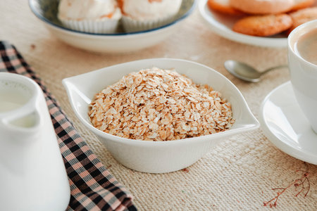 some rolled oats in a white ceramic bowl placed on a table set with a beige tablecloth next to a creamer with some plant milk and some plates with cookiesの写真素材