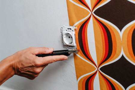 closeup of a caucasian man cutting the edges of a geometric patterned wallpaper around a socket on the wallの写真素材