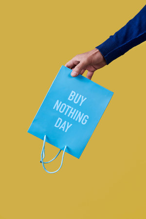 a man has a blue shopping paper bag, with the text buy nothing day written in it, upside-down in his hand, on a yellow backgroundの写真素材