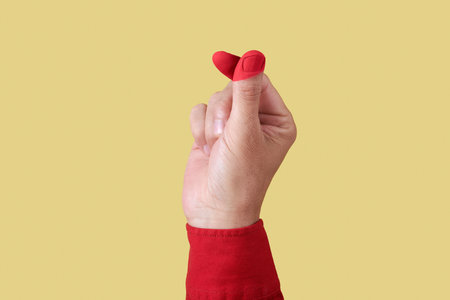 a man, wearing a red shirt, does the finger heart gesture, with his fingers painted red to form also a red heart, on a yellow backgroundの写真素材
