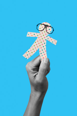 the hand of a man in black and white holding a paper man doll, wearing eyeglasses, on a blue background, as a prank for the innocents day, a feast held in some countries equivalent to april fools dayの写真素材