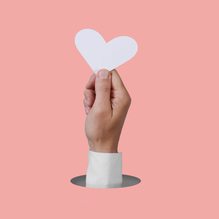the hand of a man holding a white heart popping out of a round hole on a pink backgroundの写真素材
