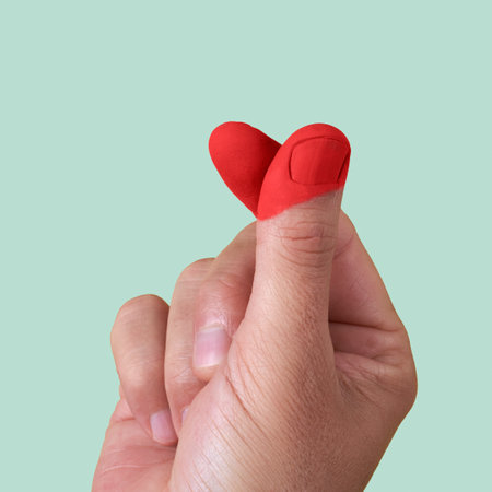 closeup of the hand of a man doing the finger heart gesture, with his fingers painted red to form also a heart, on a pale green backgroundの写真素材