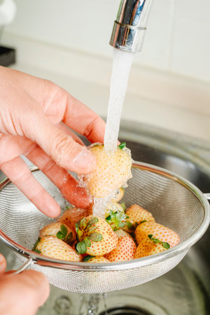 closeup of a man rinsing some white strawberries placed in a colander under the running water of the kitchen tapの写真素材