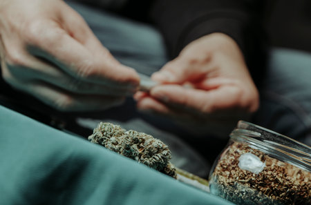 closeup of a man rolling a joint sitting at a table where there is some cannabis buds and a jar with some rolling tobaccoの写真素材