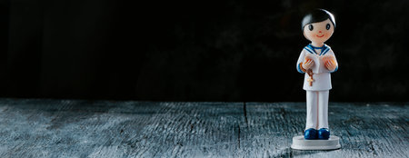 a figurine of a first communion boy, in a white sailor suit, wearing a rosary and a bible, on a rustic gray wooden surface, in a panoramic format to use as web bannerの写真素材