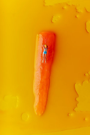 high angle view of a miniature blonde woman, wearing a blue swimsuit, lying face down on an orange freeze pop which is melting on an orangish yellow backgroundの写真素材