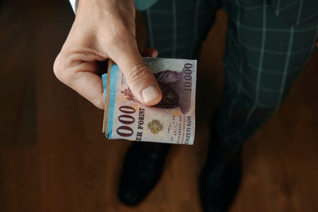 a businessman, wearing an elegant gray suit, offers a wad of hungarian forint notes to the observer, standing on a wooden floor indoorsの写真素材