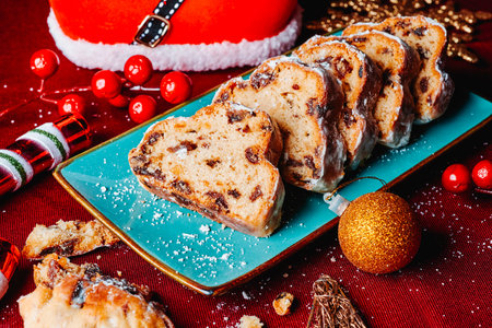a green plate with a sliced christmas stollen on a table set with a red tablecloth, next to some christmas ornaments, such as an oranment in the shape of a candy cane, a christmas ball or a starの写真素材
