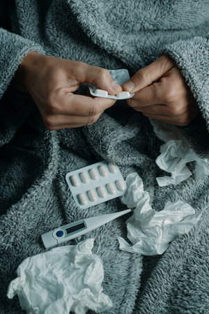 an ill man at home, wrapped in a fluffy gray house robe, is about to take a pill, surrounded by used tissuesの写真素材