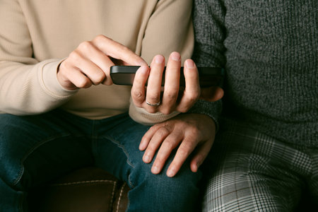 closeup of two men wearing casual clothes, sitting side by side on a brown sofa, and one of them...