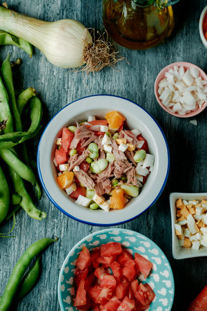 high angle view of a broad bean salad typical of andalusia, spain, made with tomato, onion, boiled egg, tuna and orange, served on a white enamel bowl, placed on a rustic gray wooden tableの写真素材