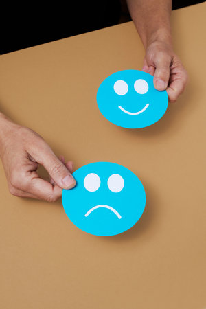closeup of a man holding a blue paper sad face and a blue paper happy face in his hands on a brown surfaceの写真素材